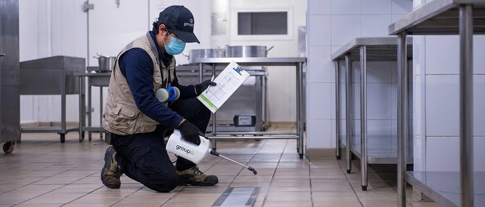 Técnico de Groupest aplicando control de plagas en una cocina industrial, con plano de puntos de control en mano y equipo de protección